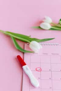 An image with a pink background, a white flower, pregnancy test (positive)and a calendar with a circled date 5th illustrating due date of baby.